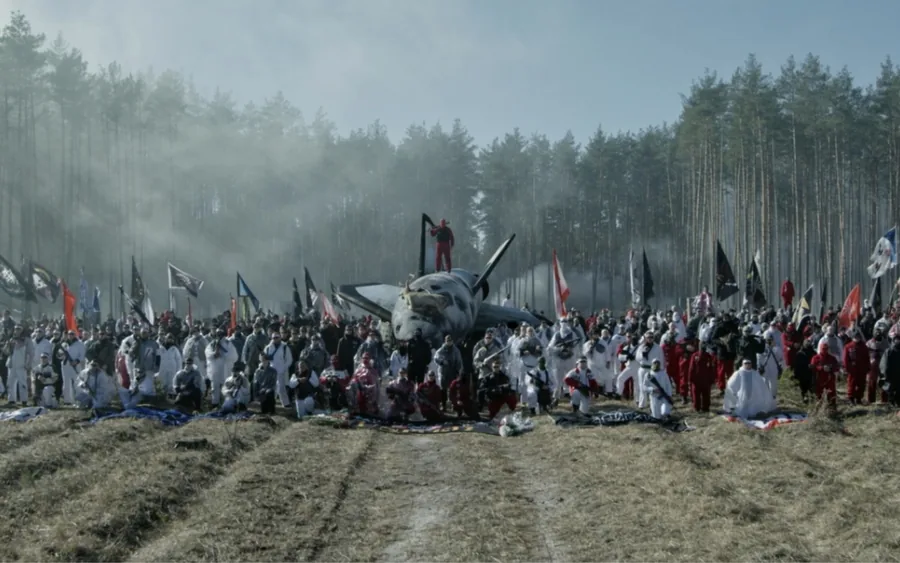 Filmstandbild aus GOD HATES SPACE von GENER8ION, gezeigt von Art Basel und Live Nation: Menschen mit Fahnen um ein Flugzeugwrack auf einer nebligen Waldlichtung